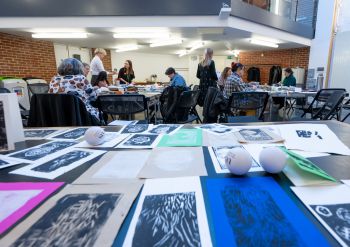 A table of lino prints with people crafting in the background