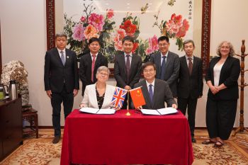 Two people sat at a red table signing an agreement with six people stanging behind them