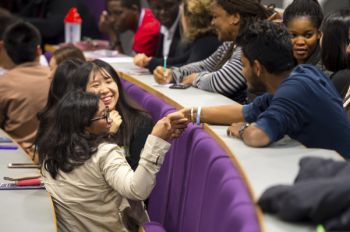 People seated in a curved lecture hall interact and converse across rows of purple chairs during a group discussion or networking event.