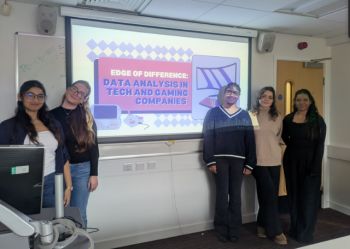 An image of four students standing next to a projector display board after their final presentation. The board reads 'Edge of Difference: Data Analysis in Tech and Gaming Companies'