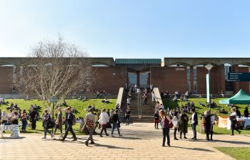Students gather and walk through Library Square on the University of Sussex campus in front of the library building on a sunny day