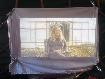 Image of a woman sitting in a greenhouse, projected onto a large fabric screen inside a dark tent.