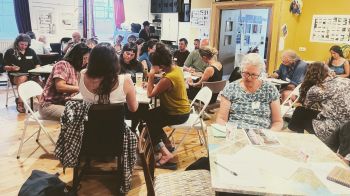 A group of people sat at tables in a bright community space, writing and drawing.
