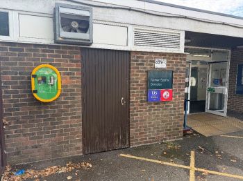A green AED box positioned on a brick wall by the main entry door to the Falmer Sports Complex.