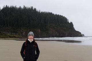 A young woman in a coat and knitted hat stands on a shoreline on the Oregon coast. Behind her we see evergreens and water.