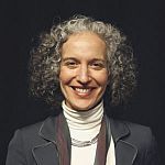 A lady with curly hair  a white top and dark jacket, smiling