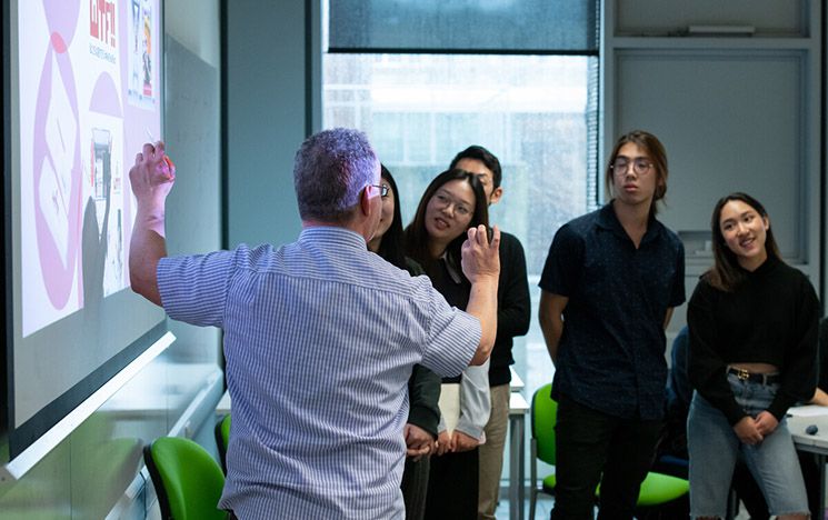 A tutor showing students a projected image on a white board. The students are stood up around the white board, looking engaged at what the teacher is showing them