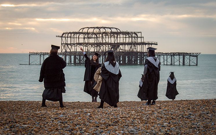 Graduates in their gowns on Brighton beach pebbles with the West Pier in the background