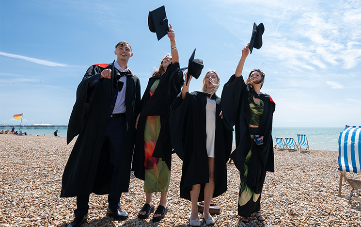 Sussex alumni on Brighton beach celebrating graduation, throwing their graduation hats in the air