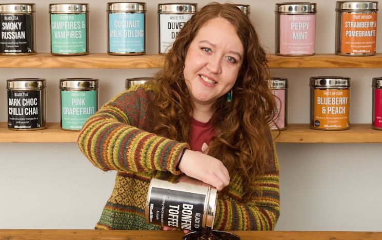 A photo of Alumnus Megan Bond standing at a table with a tin of tea leaves from Bird and Blend co.