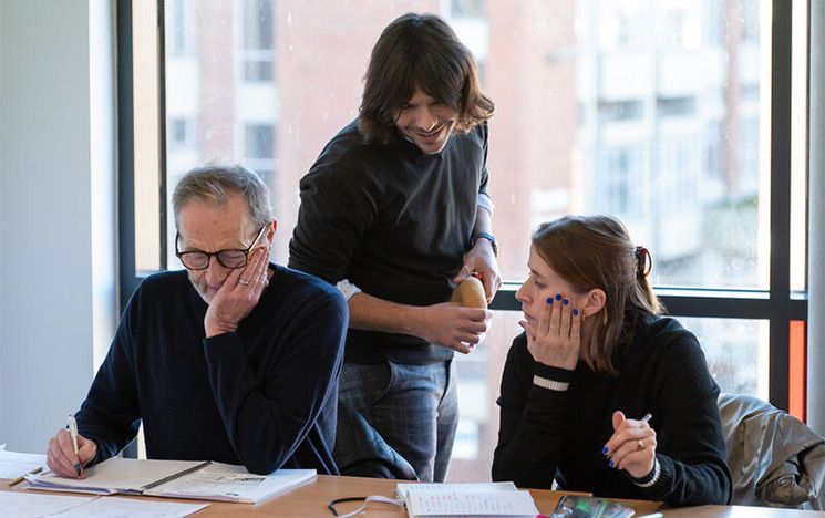 Two students study together at a table, while a tutor stands behind them observing their work