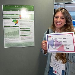 Marcela smiles directly into the camera holding her certificate and stands to the side of heraward-winning wall-mounted research poster