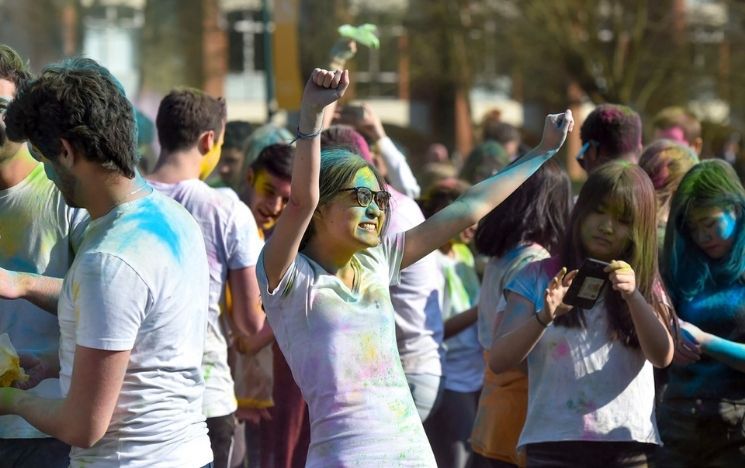 Students in white t-shirts and covered in colourful powder celebrate Holi in Library square. On the foreground, a student stretches her arms with delight