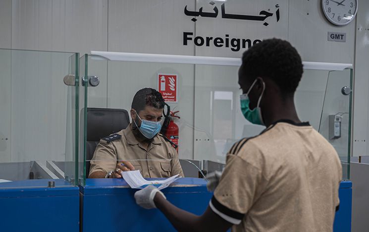 Pre-departure process, Libya, a man hands papers to a border official on the other side of the desk; a sign on the wall says 'foriegners'