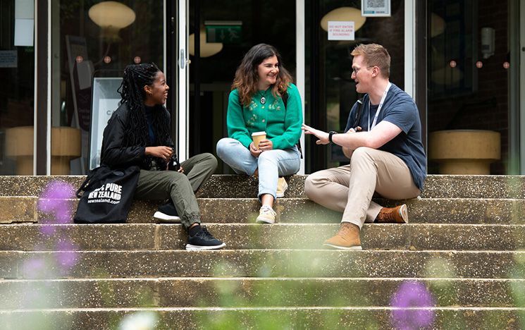 Postgraduate students sit on the steps outside a building on campus.