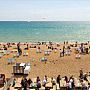 Photo of Brighton seafront and seaside, showing the pebbled beach with lots of deck chairs and people enjoying the sun, some eating fish and chips on picnic benches.