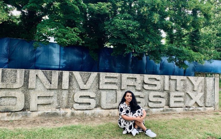 A student sits on the grass next to a stone with the words 'University of Sussex' carved into it