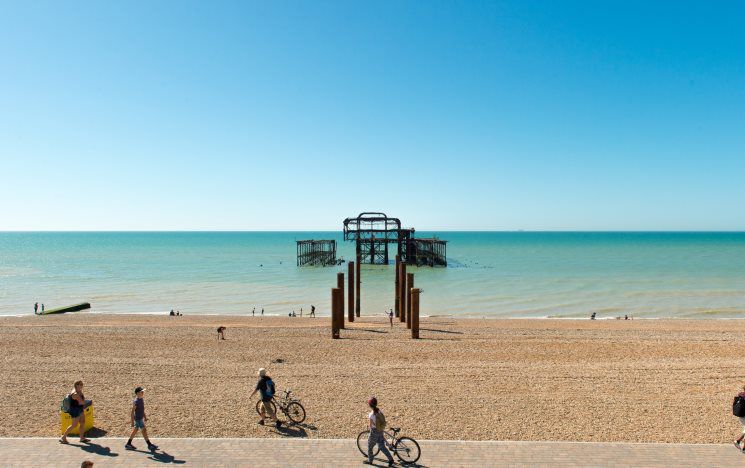 A burnt down pier on a very sunny day with a mostly empty pebble beach and blue sea