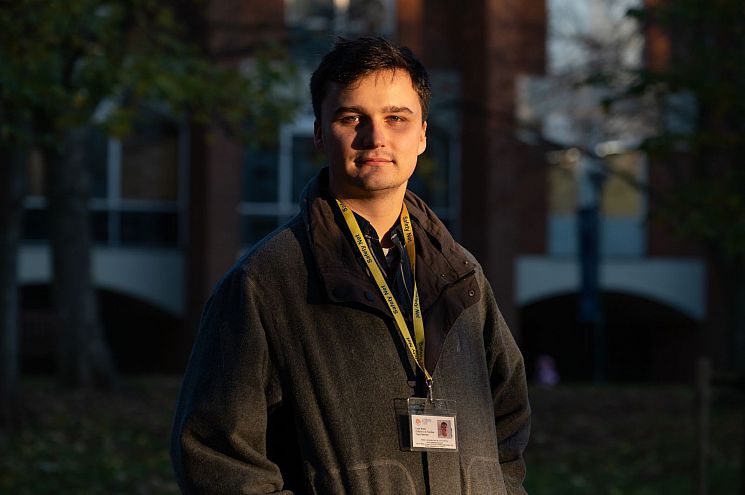 Psychology graduate Luke Bunn stands in front of red-brick buildings on campus, wearing his 'Safety Net' lanyard.