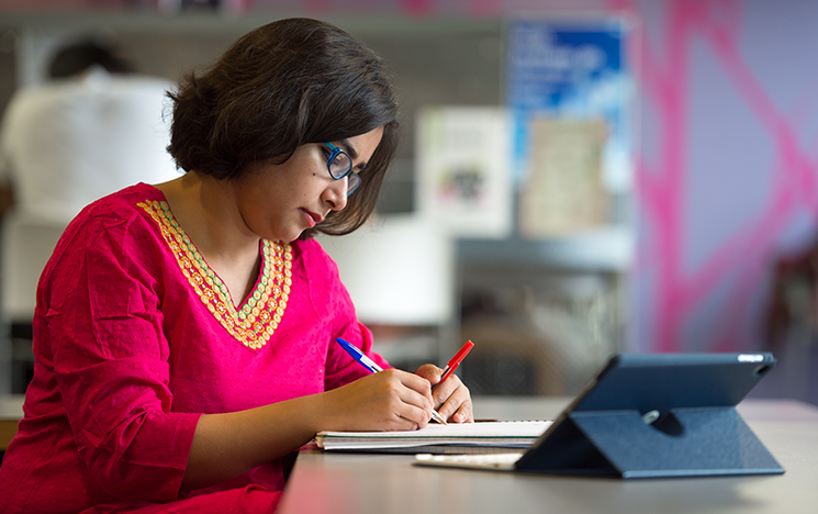 A student in a pink top leaning over a table writing on a pad with an ipad propped up on the table in front of her.
