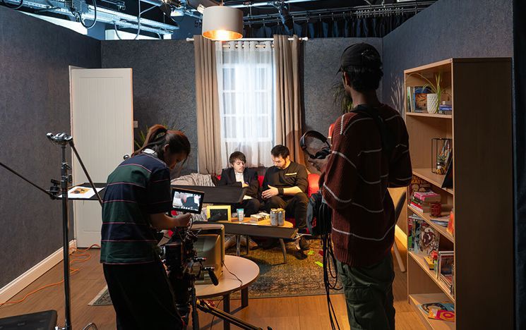 A group of students on a film set of a living room, including a camera operator and someone giving notes to an actor on a sofa