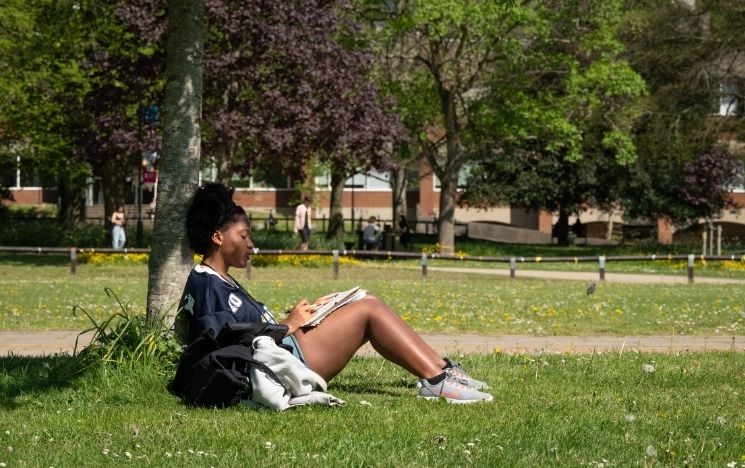A girl sits with her back against a tree in library square, writing notes in a book on a sunny day