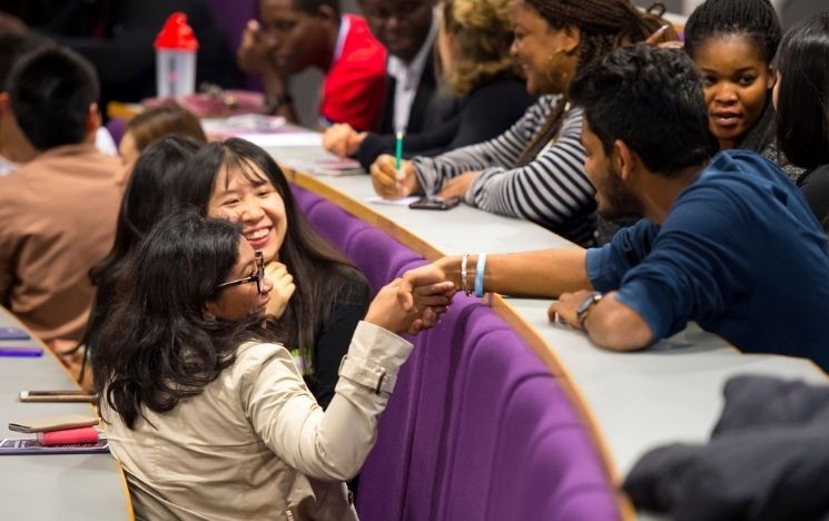 A group of students sat in a lecture theatre, turning around and shaking hands and chatting with the students behind them