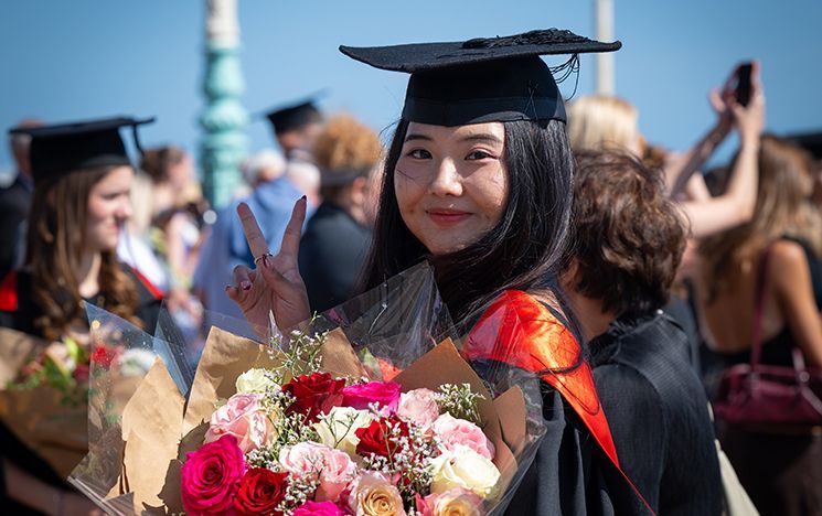 A graduate holding a large flower bouquet making a peace sign.