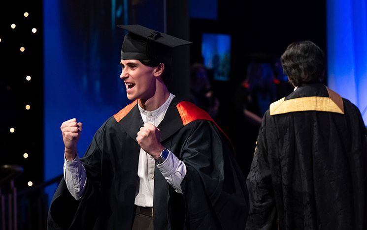 A graduate in gown raises both fists in celebration on a lit stage.