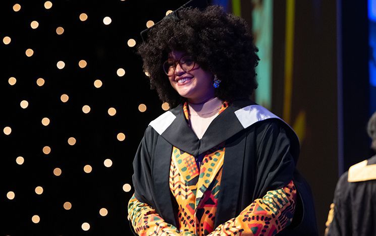 A graduate in gown with colourful patterned clothing stands on a lit stage.