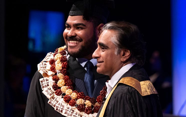 A graduate wearing a flower garland stands beside the Chancellor on stage.