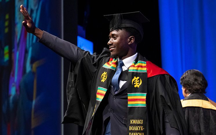 A graduate wearing a patterned stole raising an arm on stage.