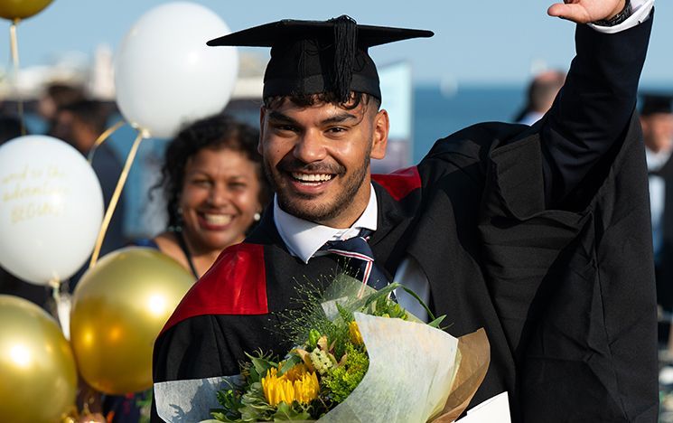 A smiling graduate holds sunflowers and raises an arm near the sea-front.