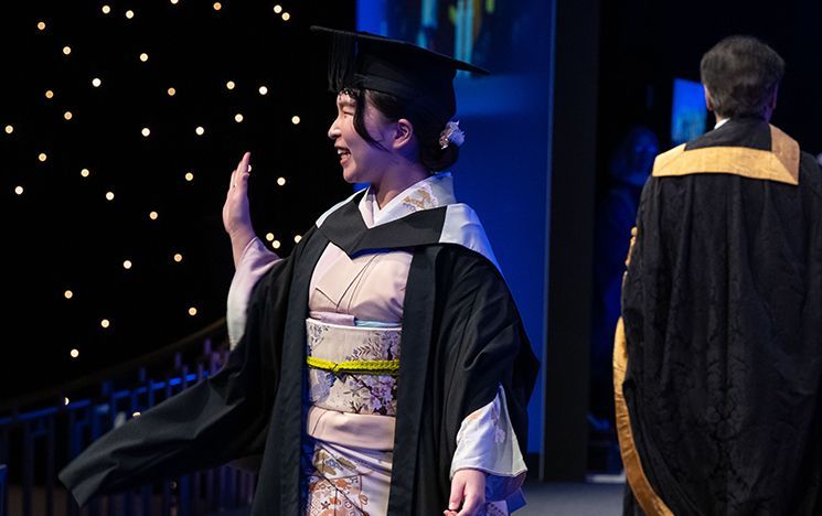 A graduate wearing a kimono under their gown walking across a blue‑lit stage.