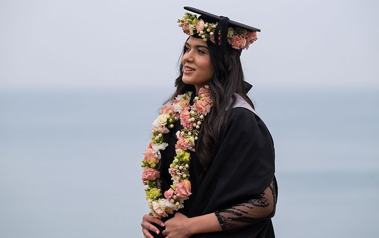 A graduate wearing a floral crown standing outdoors near the sea.