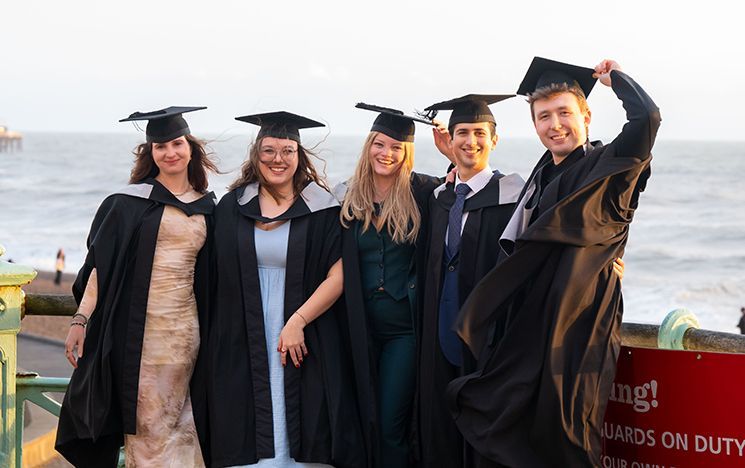 Five graduates standing together near a seaside railing at sunset.