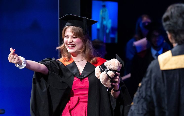A graduate in a pink outfit under their gown holding a teddy bear on stage.