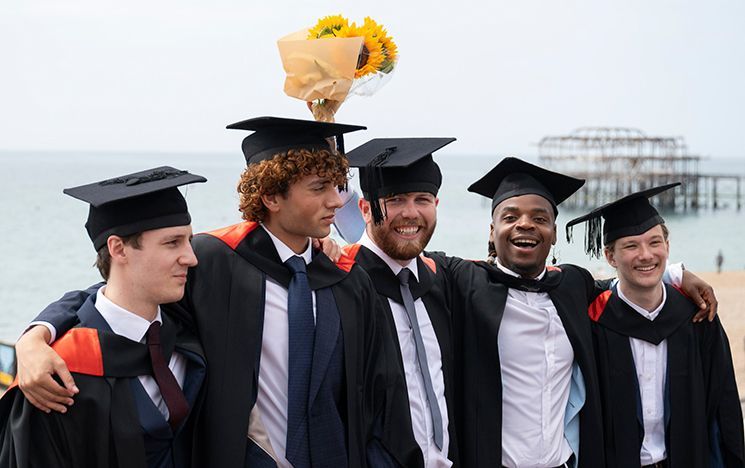 Five graduates stand on Brighton beach with the old pier behind them.