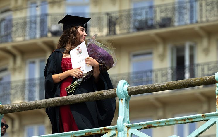 A graduate with a bouquet of flowers and certificate in hand standing behind blue railings.