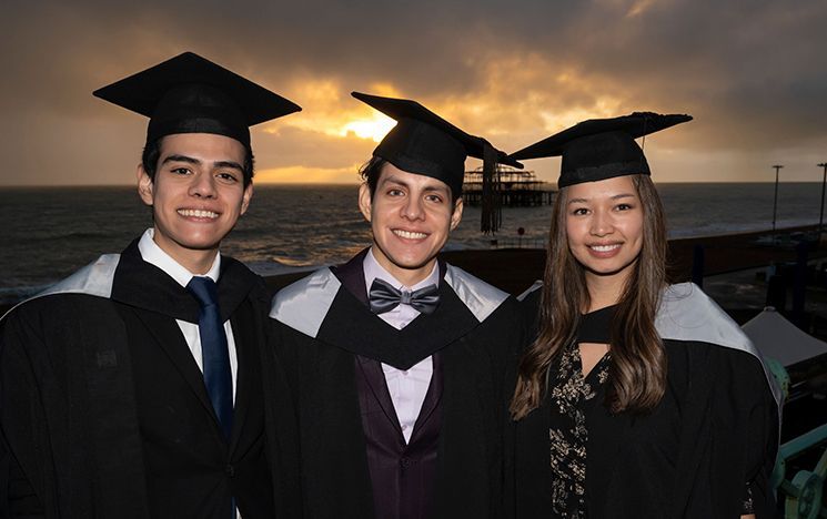 Three graduates in robes smiling in front of a sunset.