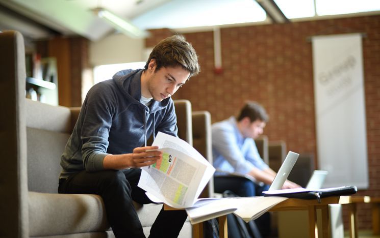 Two students sat on Library sofas, one reading a journal article, the other using a laptop