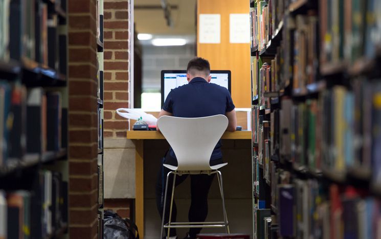 Student sat at computer between two rows of shelves