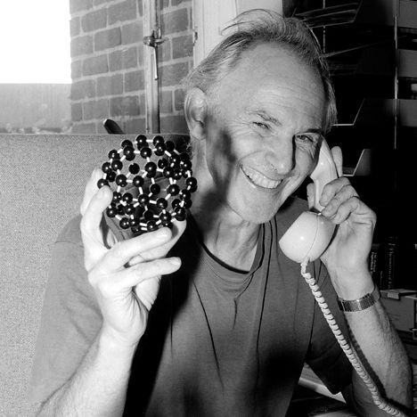 Black and white photo of Professor Harry Kroto holding up a model of a 'buckyball' while using the phone.