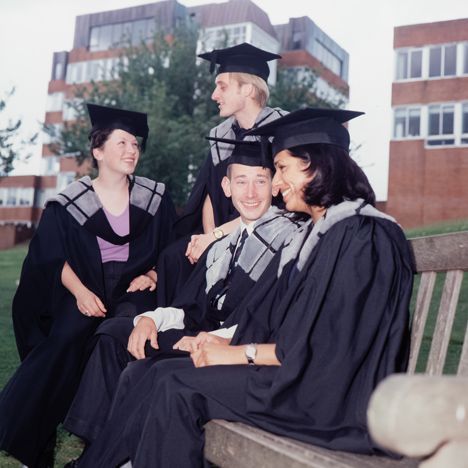 A group of students sit on a bench on campus, wearing older graduation gowns and caps.