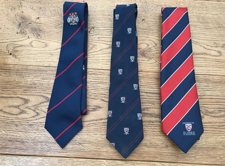 Three University of Sussex ties. One is navy blue with the crest and thin, red diagonal lines. Another is navy with smaller crests in a pattern and the last is alternating thick navy and red diagonal stripes.