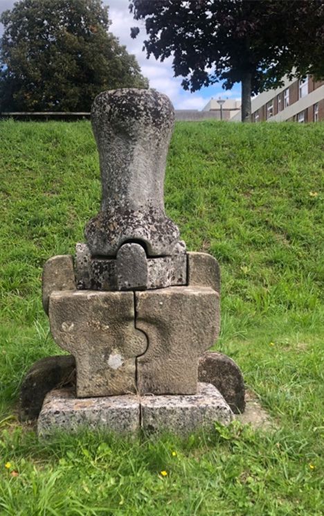 A squat stone sculpture, square at the bottom and with a vase-like shape on top, sits on the grass outside Sussex House.