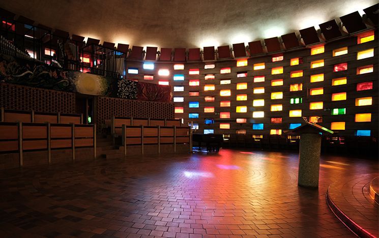 Empty interior of the Meeting House, showing the curvature in the walls of the building and with sunlight streaming through the multiple, multicoloured windows.