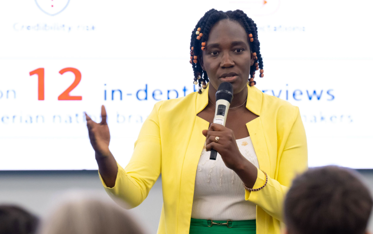 A speaker gesturing with one hand and speaking into a microphone while wearing a bright yellow blazer during a presentation.