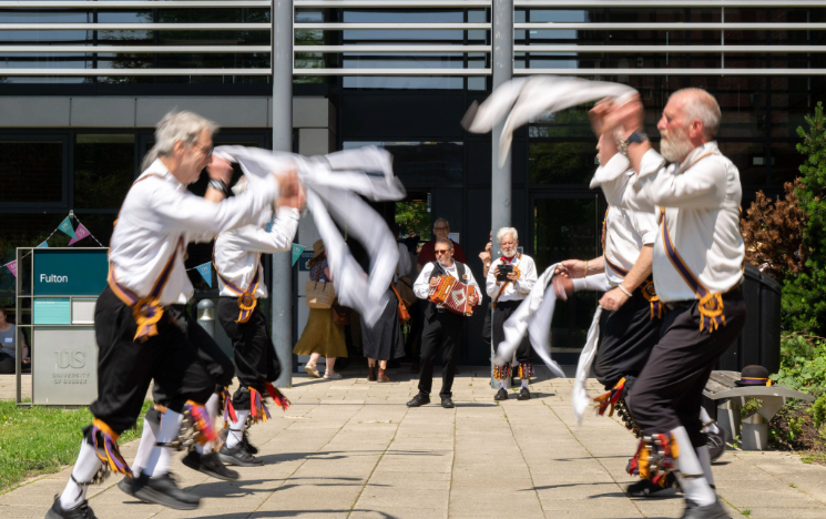 Morris dancers in traditional white shirts, dark trousers, and bells perform outdoors with waving white handkerchiefs to music played by an accordionist.
