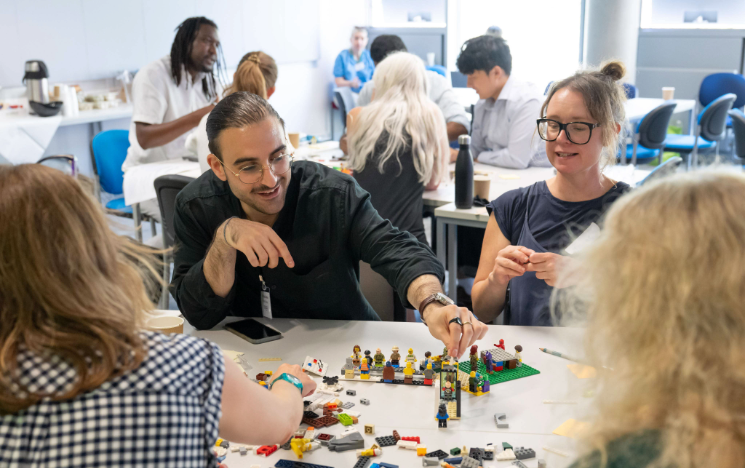 A group of people collaborate around a table covered with colorful lego building blocks, assembling a small model together in a bright seminar room.